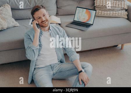 Young man talks on smartphone beside laptop with charts while working from home Stock Photo