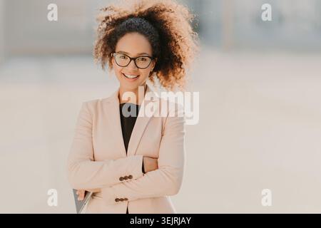 Smiling businesswoman with glasses standing arms crossed outdoors in blazer Stock Photo