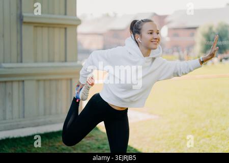Young woman stretches her leg while wearing wireless earbuds before an outdoor run in a city park Stock Photo