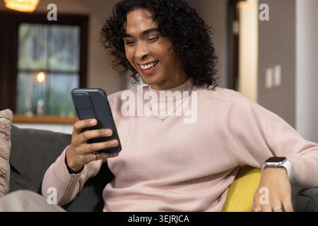 Non-binary adult sitting on gray sofa at home, holding smartphone and smiling, wearing smartwatch. Phone, device, individual, li Stock Photo