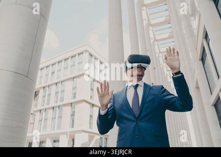 Businessman wearing virtual reality headset and gesturing outside modern office building Stock Photo