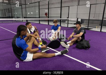 Diverse male friends sitting on purple padel court near net holding rackets and balls. Players, group, athletic, facility, equip Stock Photo
