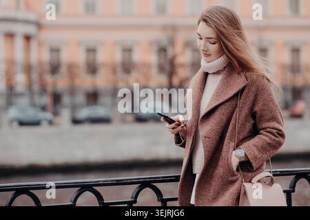 Young woman in brown coat uses smartphone near riverside railing in city Stock Photo