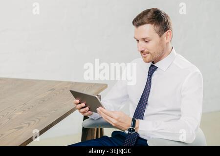 Businessman in white shirt and tie uses tablet while seated at modern office table Stock Photo