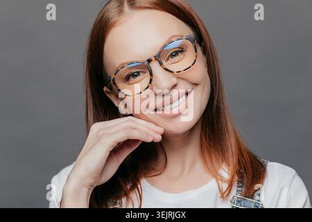 Smiling young red-haired woman wearing eyeglasses poses for close-up portrait against gray studio wall Stock Photo