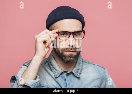 Bearded man in eyeglasses and black beanie adjusting frames in close studio portrait on pink backdrop Stock Photo