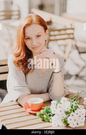 Red-haired young woman sits at outdoor cafe table with coffee cup and white flower bouquet Stock Photo
