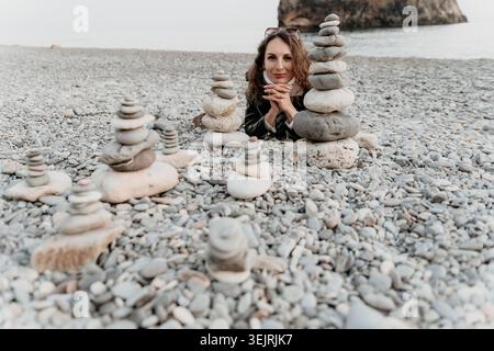 Woman beach cairns posing behind stacks of balancing stones on a pebble shore during a peaceful cloudy day Stock Photo