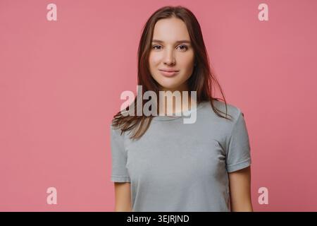 Young brunette woman in gray t-shirt looks at camera against pink studio backdrop Stock Photo