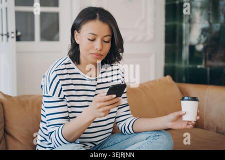 Young woman using smartphone and holding takeaway coffee while sitting on sofa at home Stock Photo