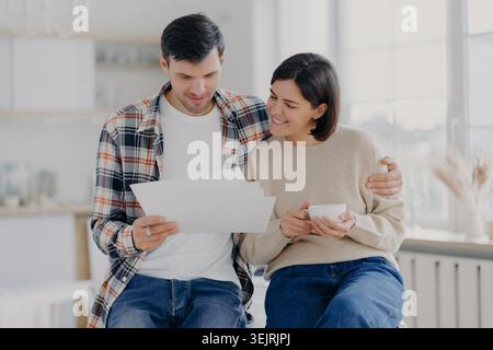 Happy young couple reads household financial documents together in bright home kitchen Stock Photo