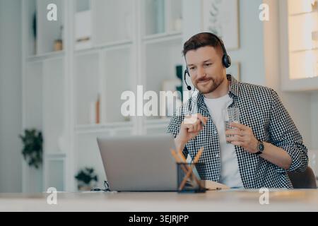Man wearing headset drinks water while working on laptop in bright home office Stock Photo