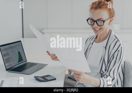 Smiling female accountant reviews financial reports with laptop and calculator at office desk Stock Photo