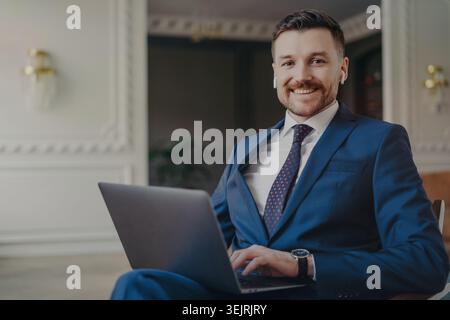 Smiling businessman in blue suit uses laptop and wireless earbuds in office lounge Stock Photo