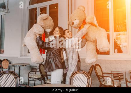 Women teddy bears friendship Two joyful friends hold massive plush toys outside a city cafe during a golden sunset Stock Photo
