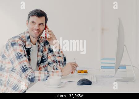 Young man talks on smartphone while working at desktop computer in bright home office Stock Photo