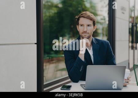 Businessman works on laptop with coffee at outdoor cafe table near window Stock Photo