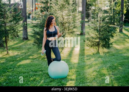 Woman in black activewear stands with exercise ball while using smartphone and headphones in park Stock Photo
