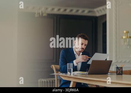 Businessman reviews documents with laptop and earbuds at office desk Stock Photo