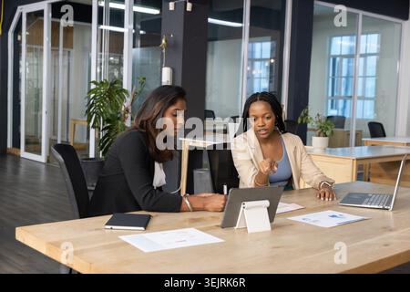 African American and Indian female team in suits discussing tablet on stand at office with charts. Coworking, collaboration, tea Stock Photo