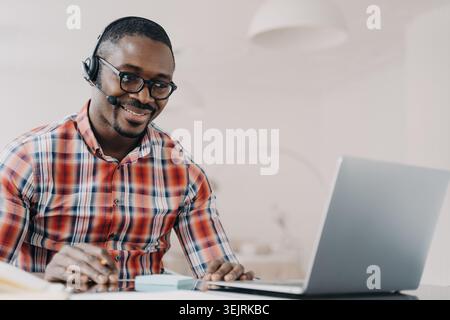 Smiling customer service representative wearing headset works on laptop at home office desk Stock Photo