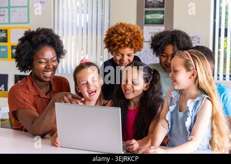 Female teacher with six children leaning and pointing at open laptop on white table in classroom. Vibrant, collaborative, learne Stock Photo
