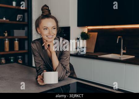 Young Woman in Satin Pajama Robe Smiling with Coffee Mug While Leaning on Counter in Modern Dark Kitchen During Morning Routine Stock Photo