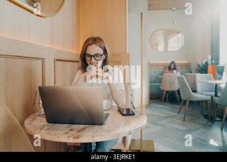 Red haired woman with glasses smiles while working on laptop in modern cafe Stock Photo