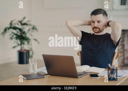 Caucasian man stretches with hands behind head while working on laptop in home office Stock Photo