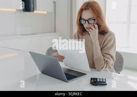 Red-haired woman with glasses reviews financial documents beside laptop and calculator at home Stock Photo