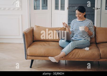 Young woman drinks coffee while working on laptop on brown sofa in bright living room Stock Photo