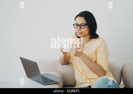 Young woman in glasses drinks coffee while working on laptop on sofa at home Stock Photo