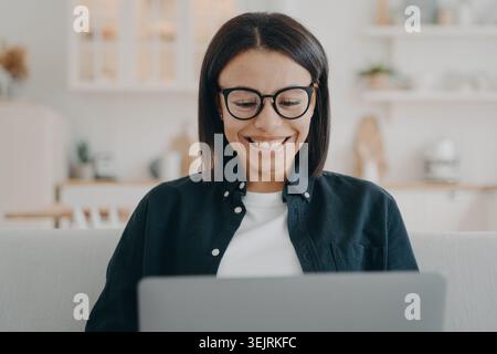 Smiling young Black woman wearing glasses works on laptop from home Stock Photo