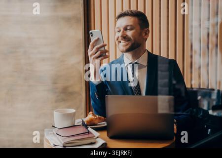 Businessman using smartphone beside laptop during coffee break in cafe by window Stock Photo