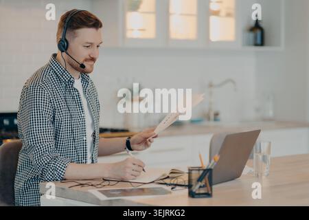 Remote support agent wearing headset reviews paperwork and writes notes beside laptop in home kitchen office Stock Photo