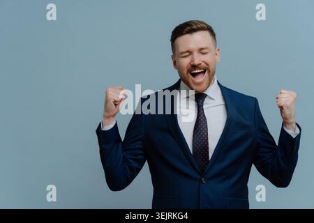 Excited businessman in blue suit celebrates success with raised fists in studio Stock Photo