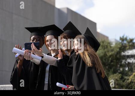 Female graduates wearing caps and gowns taking group selfie on campus, holding phone and diplomas. Students, celebration, milest Stock Photo