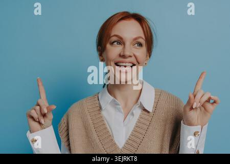 Smiling redhead woman points upward with both fingers against blue studio wall Stock Photo