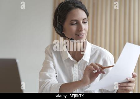 Female customer support specialist wearing headset reviews printed document while working with laptop in office Stock Photo