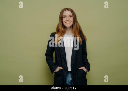 Young redhead woman in dark blazer smiles with hands in pockets against green studio wall Stock Photo