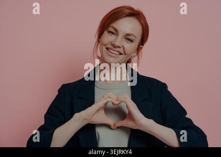 Smiling red haired woman makes heart shape with hands against pink studio backdrop Stock Photo