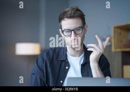 Young man with glasses gestures while using laptop in home office Stock Photo