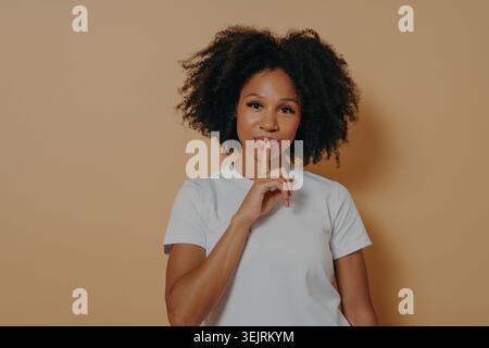 Young Black woman makes hush gesture with finger on lips in beige studio portrait Stock Photo