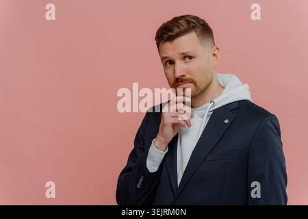 Man in navy blazer and gray hoodie touches chin while posing against pink studio wall Stock Photo
