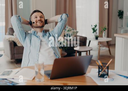 Young man wearing headset leaning back with hands behind head at home office desk with laptop, relaxing after work call Stock Photo
