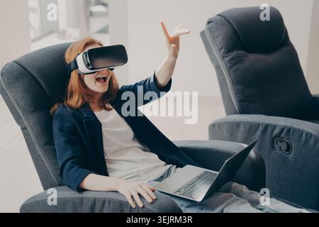 Young red-haired woman in blazer wearing VR headset with laptop on lap gesturing excitedly in modern lounge chair Stock Photo