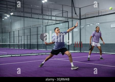 Adult men playing padel, foreground player lunging with racket, wearing sportswear on purple court. Athletes, paddle, doubles, net, glass, walls, ball Stock Photo