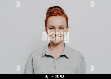 Young red-haired woman with updo smiling confidently at camera wearing casual plaid shirt against light gray background Stock Photo