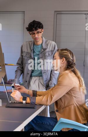Coworkers working at office table, woman leaning over laptop with stylus, man with glasses watching. Duo, boardroom, flipchart, tablet, smartwatch, co Stock Photo