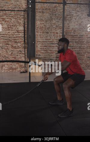 African American man using battle ropes, squatting in gym with rig, wearing red shirt, copy space Stock Photo